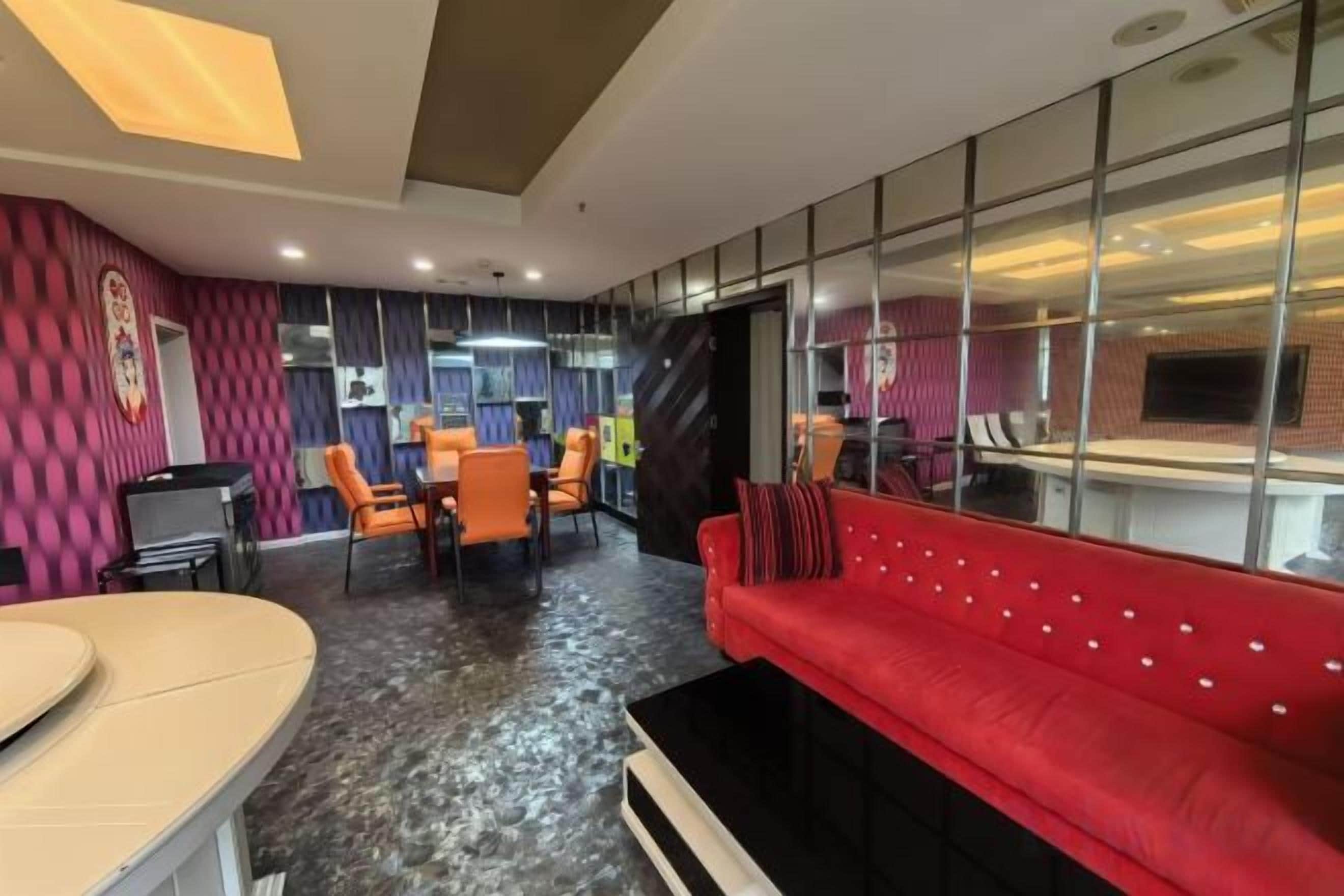 A modern hotel lobby with a red sofa, orange chairs, a dining area, and a large mirror reflecting the space.