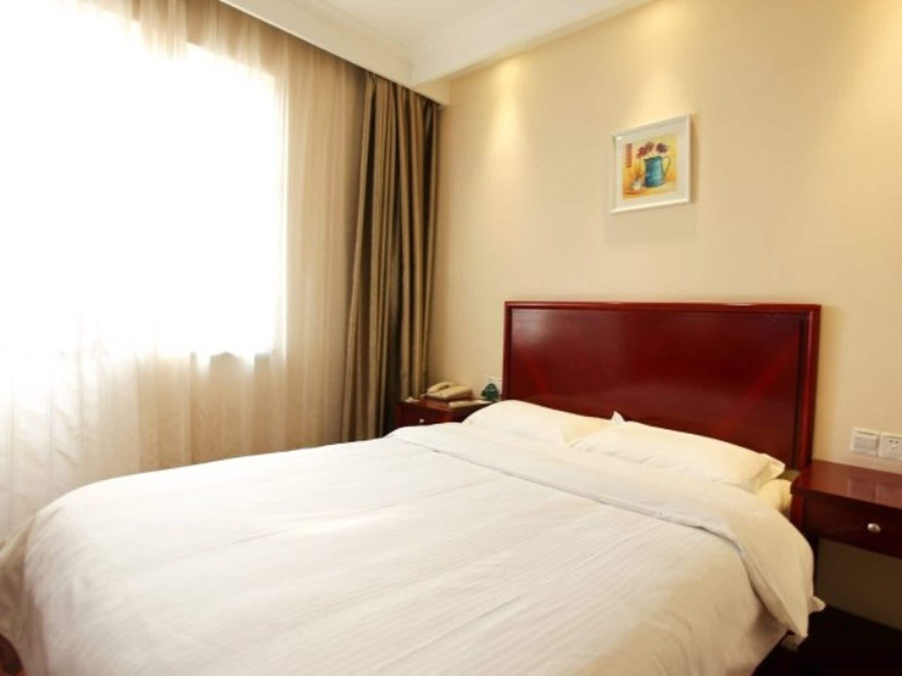 A hotel room with a large bed, a wooden headboard, a bedside table with a lamp, and a window with curtains.