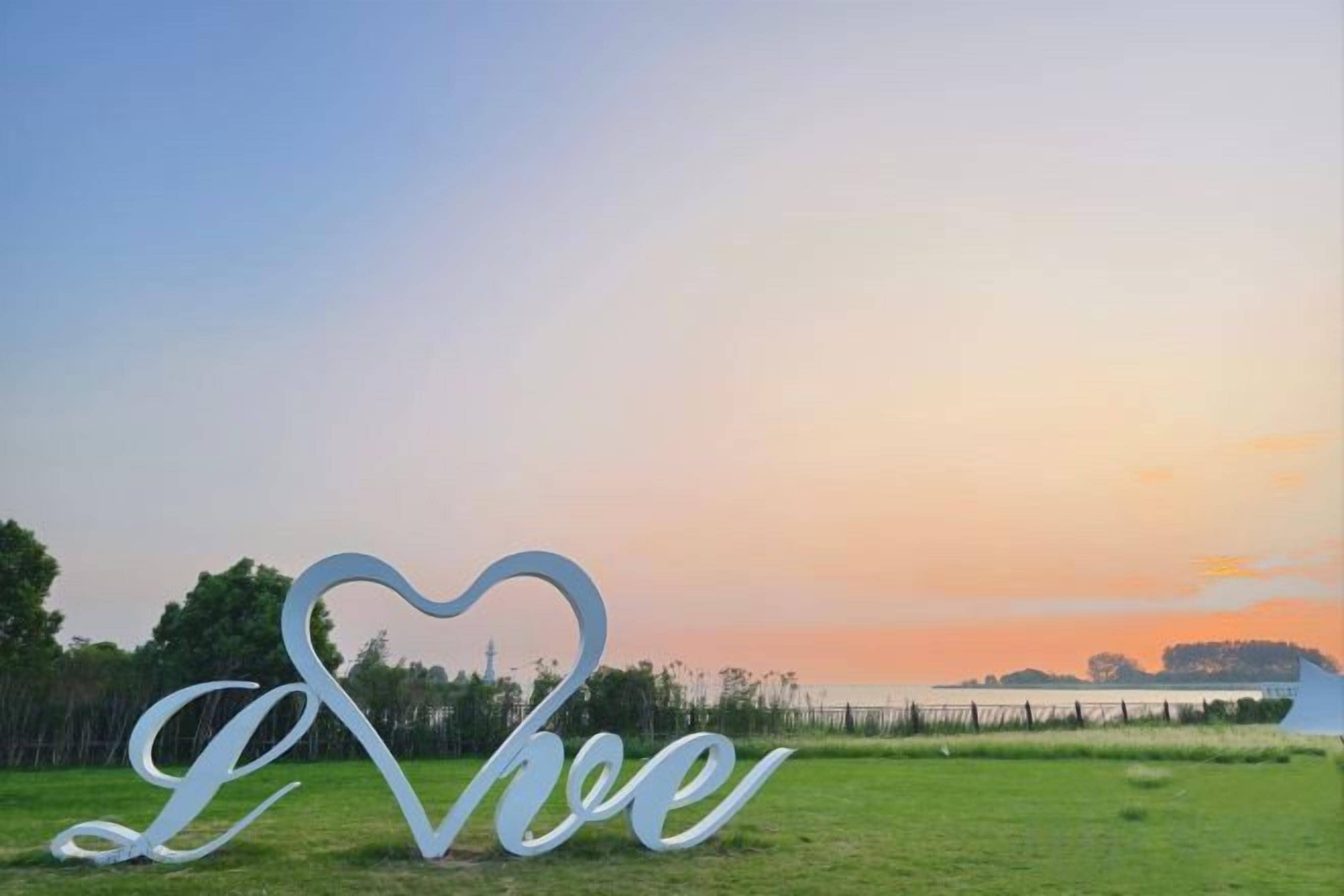Un letrero con forma de corazón que dice 'I Love' en un campo con un atardecer de fondo.