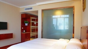 A bedroom with a bed, a TV mounted on the wall, a wooden wardrobe, and a large window with curtains.