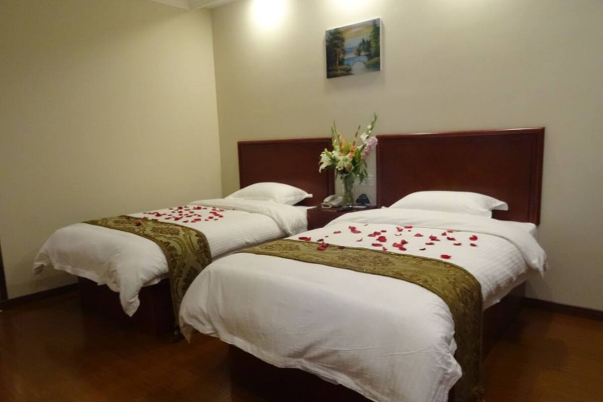 Een hotelkamer met twee enkele bedden, een houten hoofdbord, een nachtkastje met een bloevas en een schilderij aan de muur.