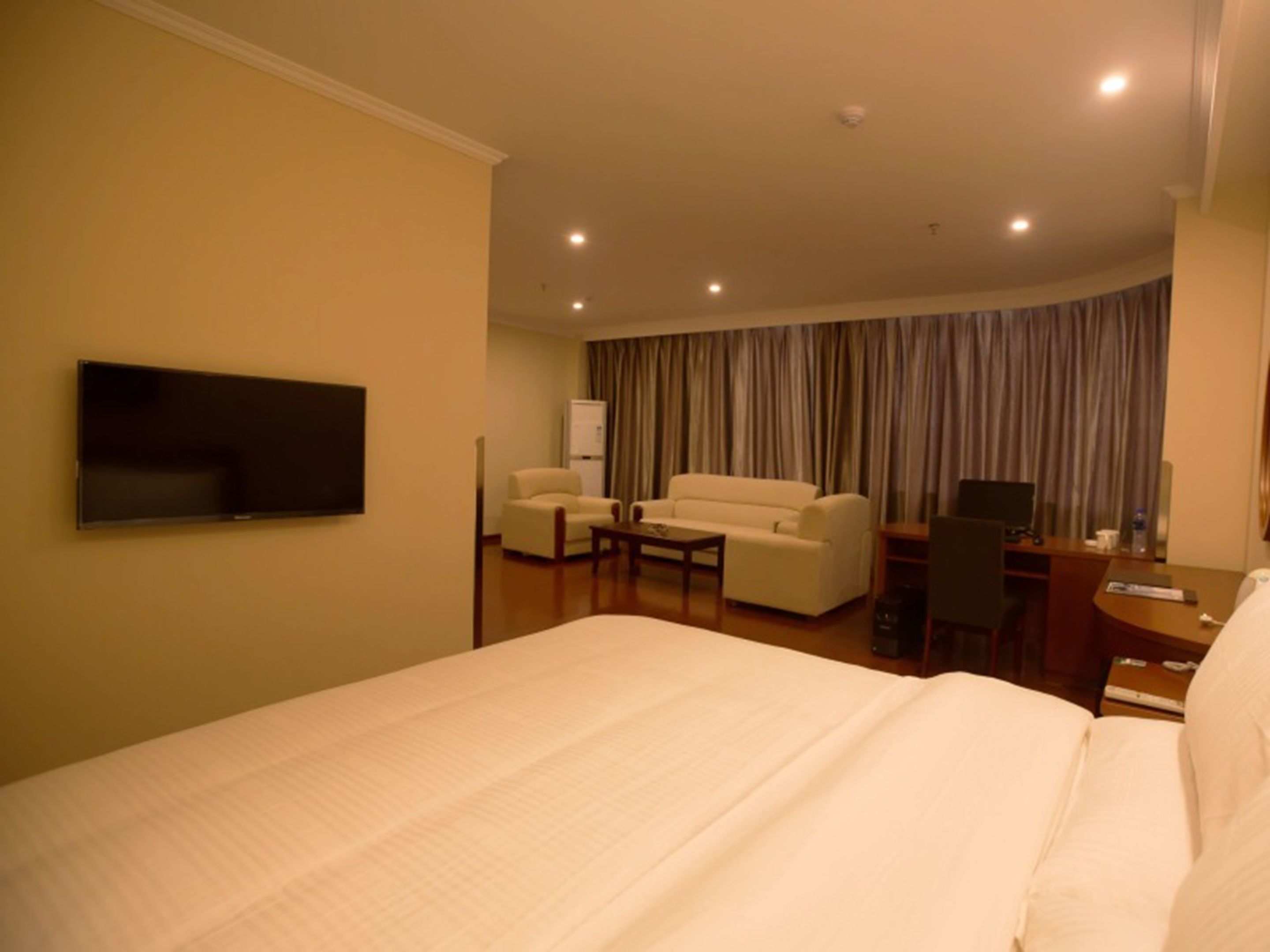 A hotel room with a large bed, a TV mounted on the wall, a desk with a chair, a sofa, and a curtain backdrop.