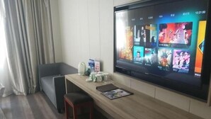 A modern hotel room with a large flat-screen TV mounted on the wall displaying various movie posters. There is a wooden bench with a magazine on it, a small table with a kettle, and a chair. - GreenTree Inn JiangSu YanCheng JianHu ShangGang Bus Station Freeway 204 Express Hotel (Yancheng)
