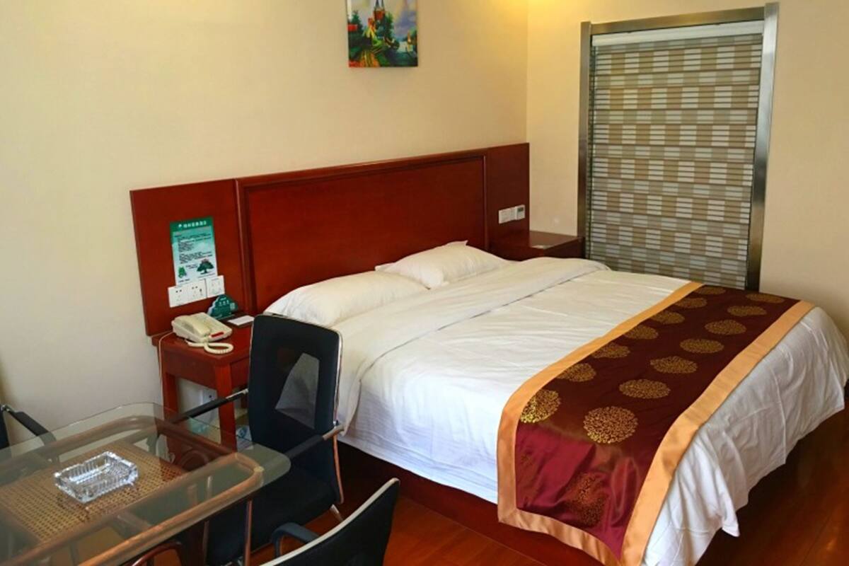 Een hotelkamer met een houten hoofdeinde, een bed met witte lakens, een glazen tafel met een stoel, een telefoon en een schilderij aan de muur.