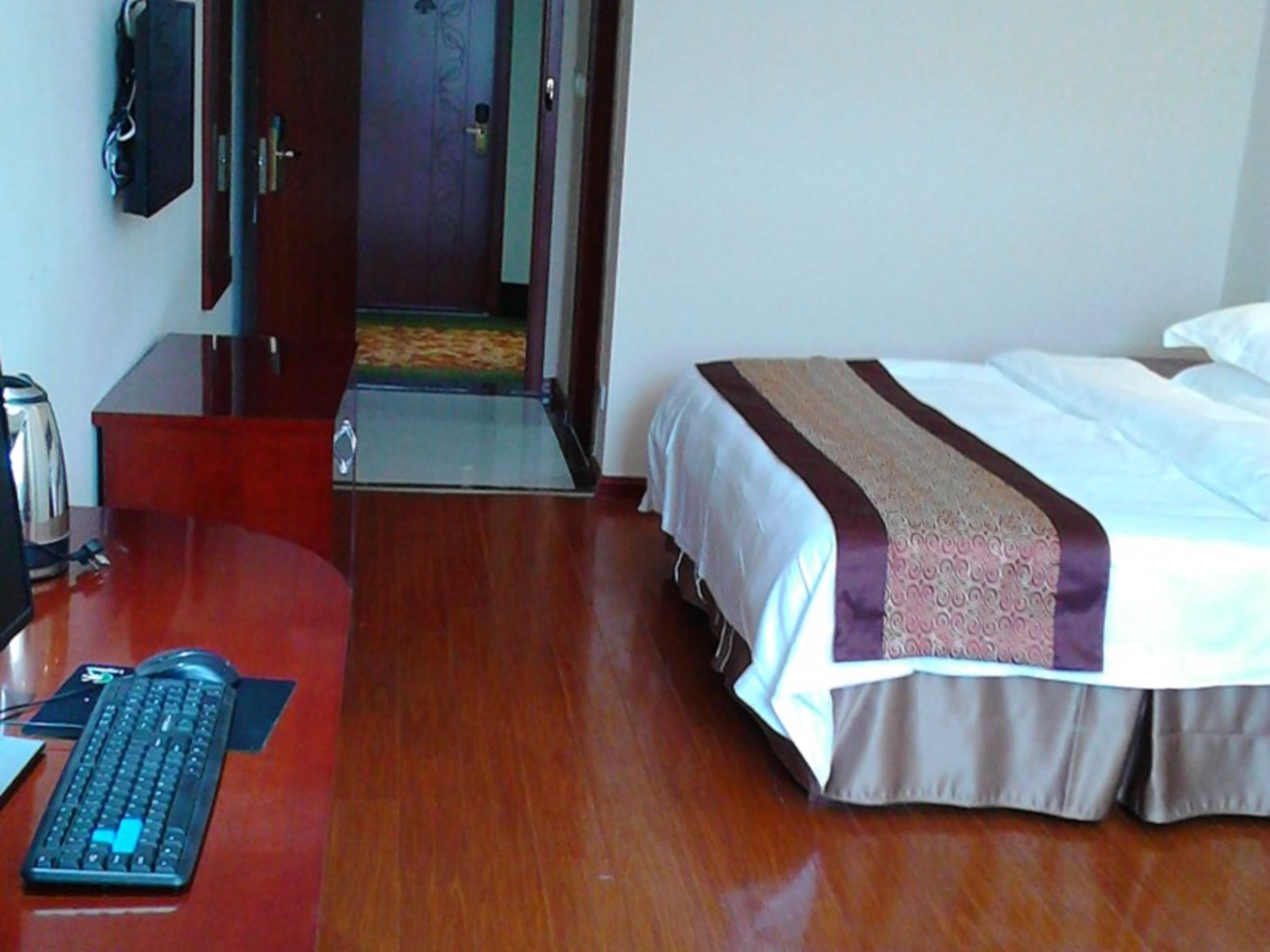 Une chambre d’hôtel avec un bureau en bois, un lit recouvert d’une courtepointe à motifs et une porte donnant sur un couloir.