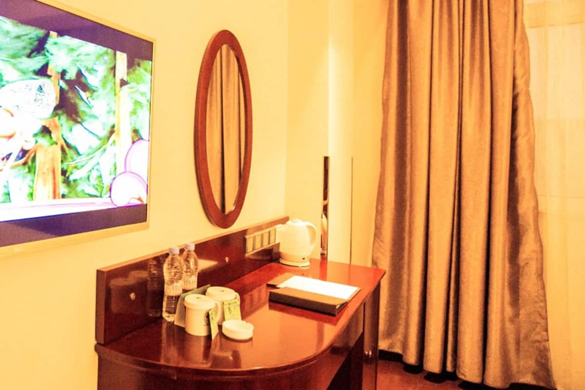 A hotel room with a wooden desk, a television mounted on the wall, a mirror, a window with curtains, and a bedside table with a lamp and a book.