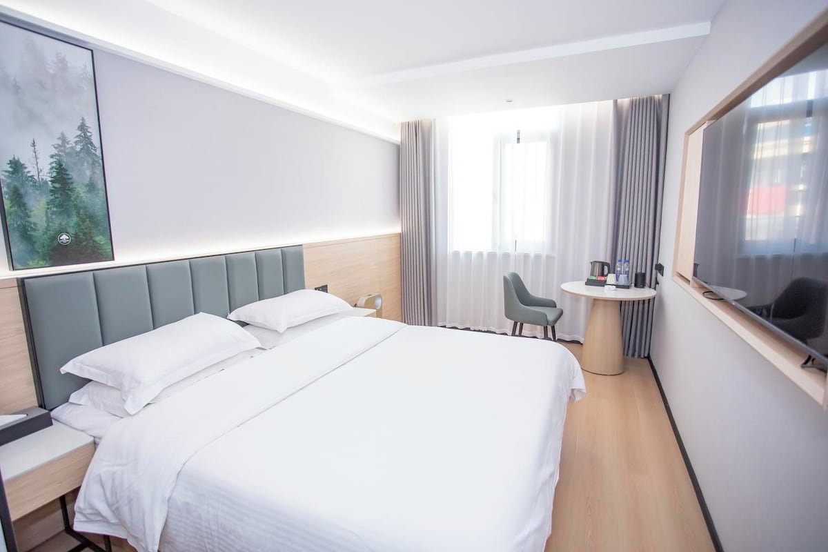 A hotel room with a large bed, a desk, a chair, a small table with a kettle, and a window with curtains.