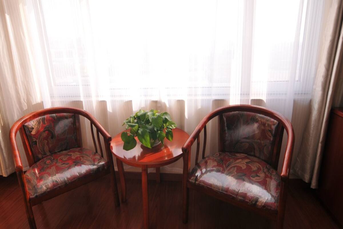 Ein Zimmer mit zwei Holzstühlen, einem kleinen runden Tisch mit einer Topfpflanze und einem großen Fenster mit transparenten Vorhängen.