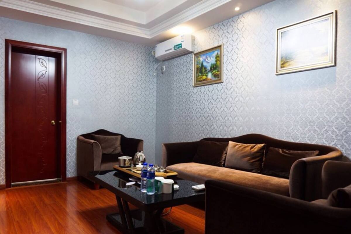 A living room with a brown sofa, a smaller armchair, a coffee table with items, a wooden floor, patterned wallpaper, a door, and an air conditioning unit.