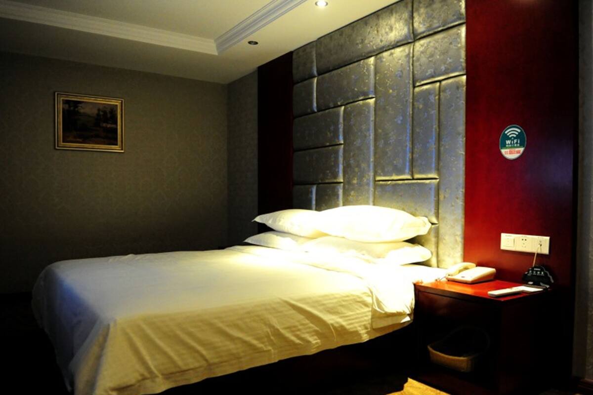 A hotel room with a bed, a nightstand with a phone and a lamp, a wall-mounted WiFi sign, and a painting on the wall.