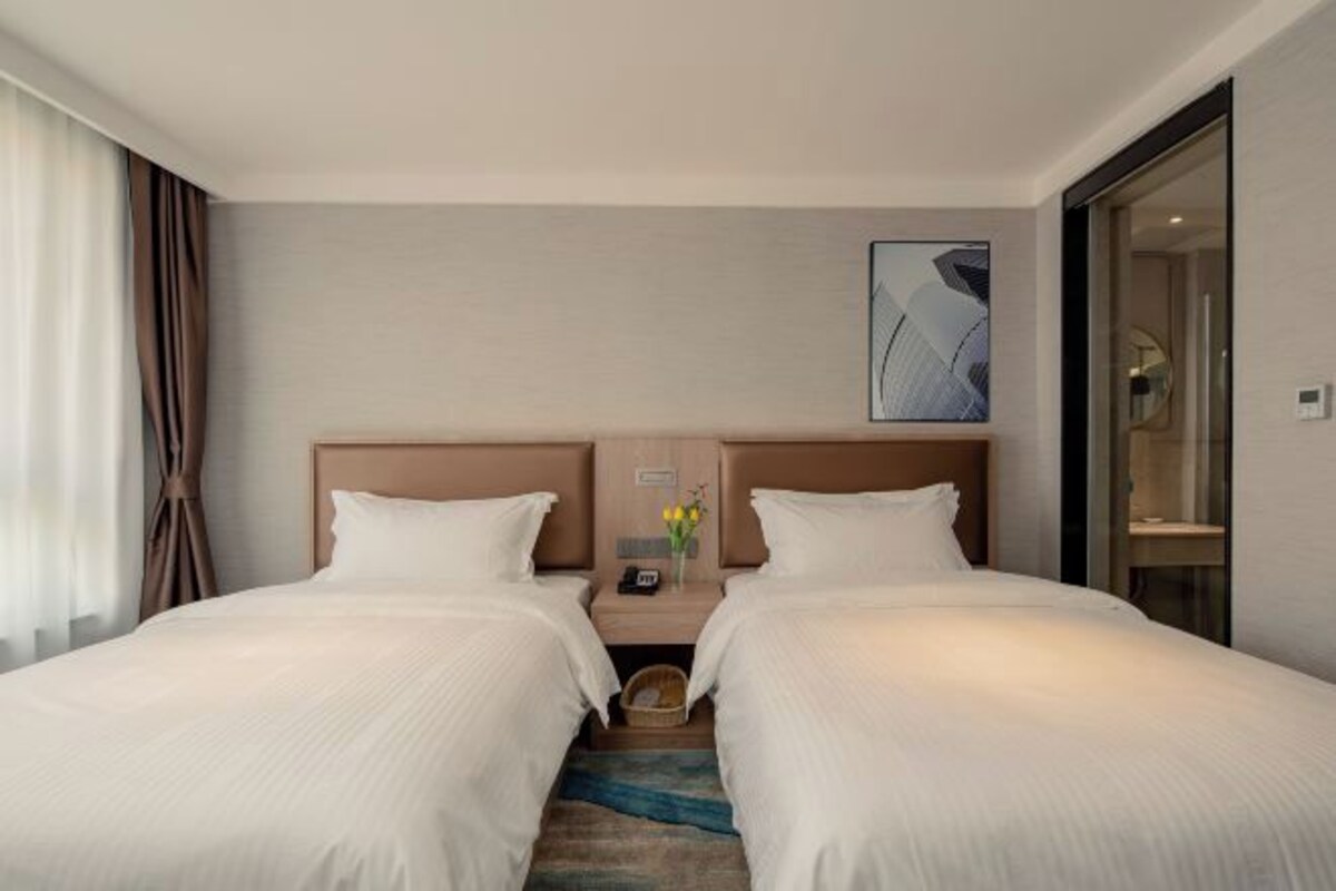 A hotel room with two beds, a nightstand with a phone and a vase of flowers, and a painting on the wall.
