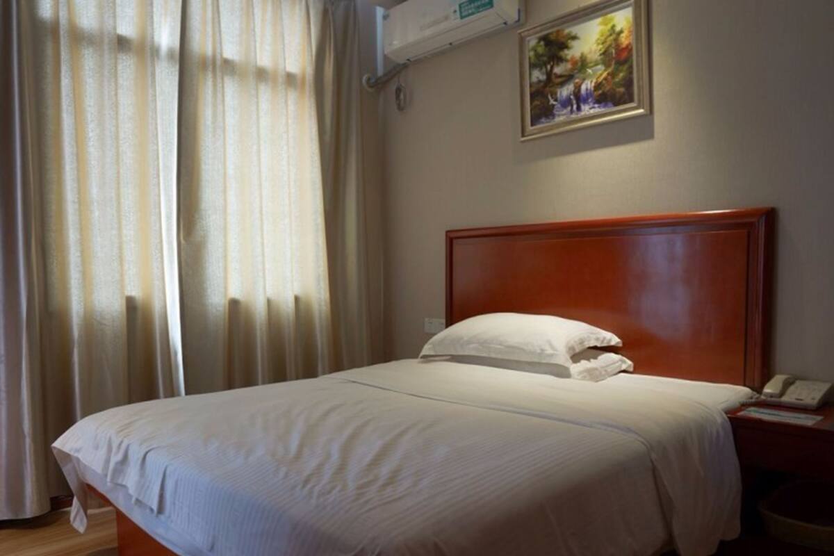 A hotel room with a wooden headboard, a white bedspread, a white pillow, a bedside table with a phone, and a painting on the wall.