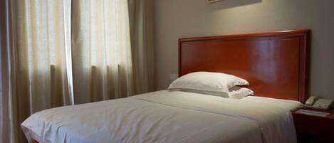 Una camera d'albergo con una testiera in legno, una coperta bianca, un cuscino bianco, un comodino con un telefono e un quadro appeso al muro.