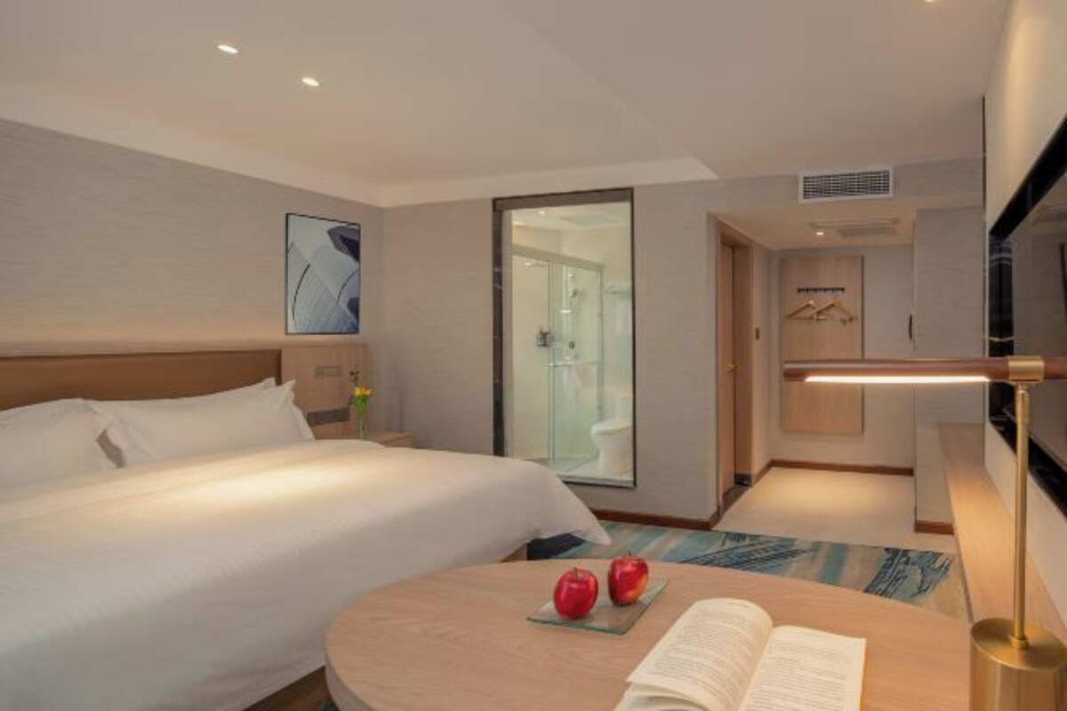 A hotel room with a bed, a bedside table with a book and apples, a desk lamp, a shower area, and a wall-mounted TV.