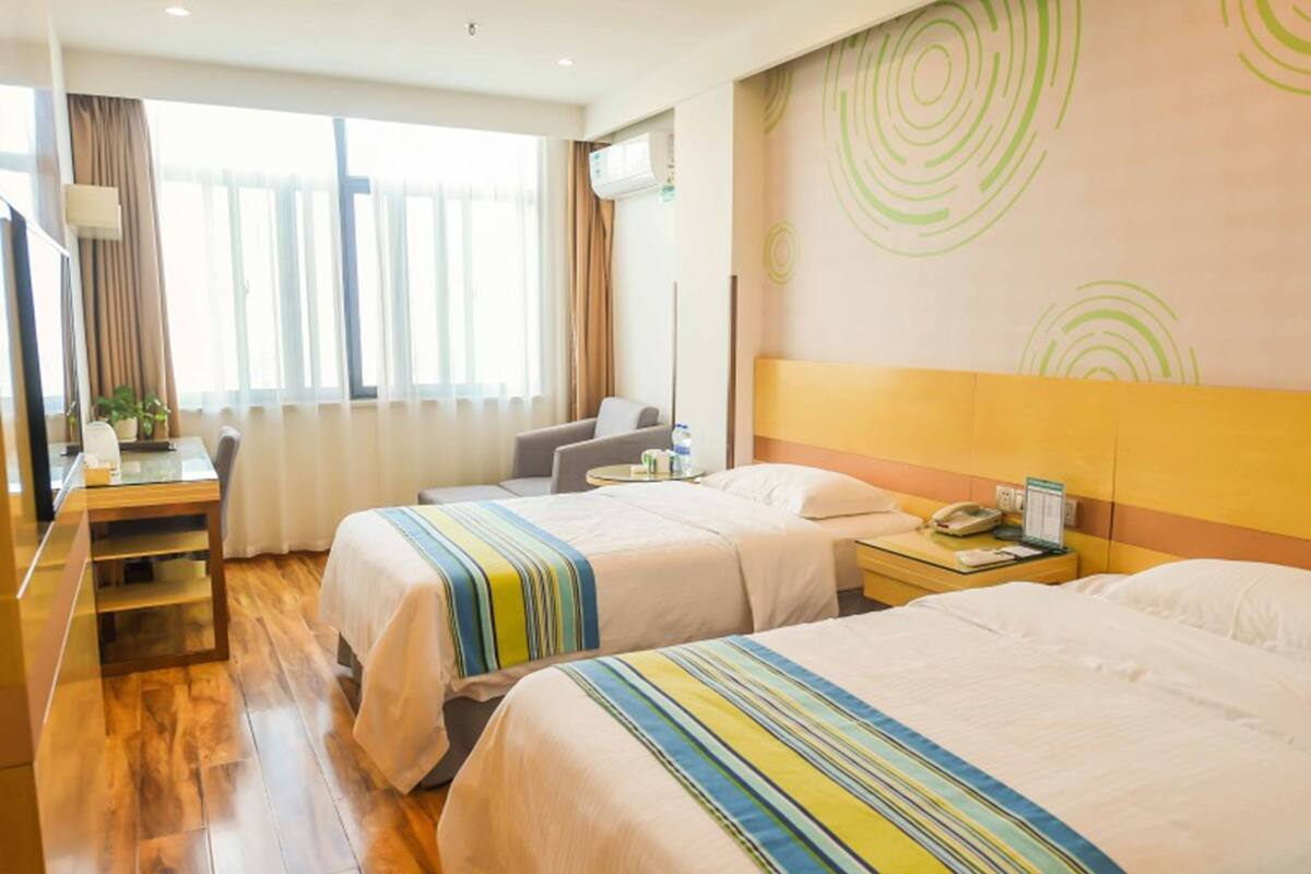 Ein Hotelzimmer mit zwei Betten, einem Schreibtisch und einem Sessel. Es gibt ein Fenster mit Vorhängen, eine Klimaanlage und eine Wand mit kreisförmigen Mustern.
