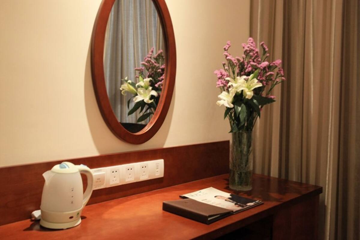 A hotel room with a wooden desk, a white electric kettle, a vase with flowers, and a mirror on the wall.