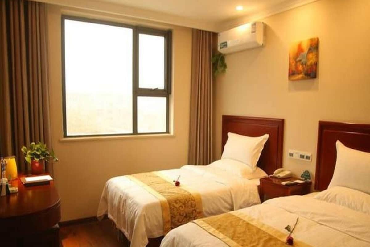 A hotel room with two beds, a desk, a window, an air conditioner, and a painting on the wall.