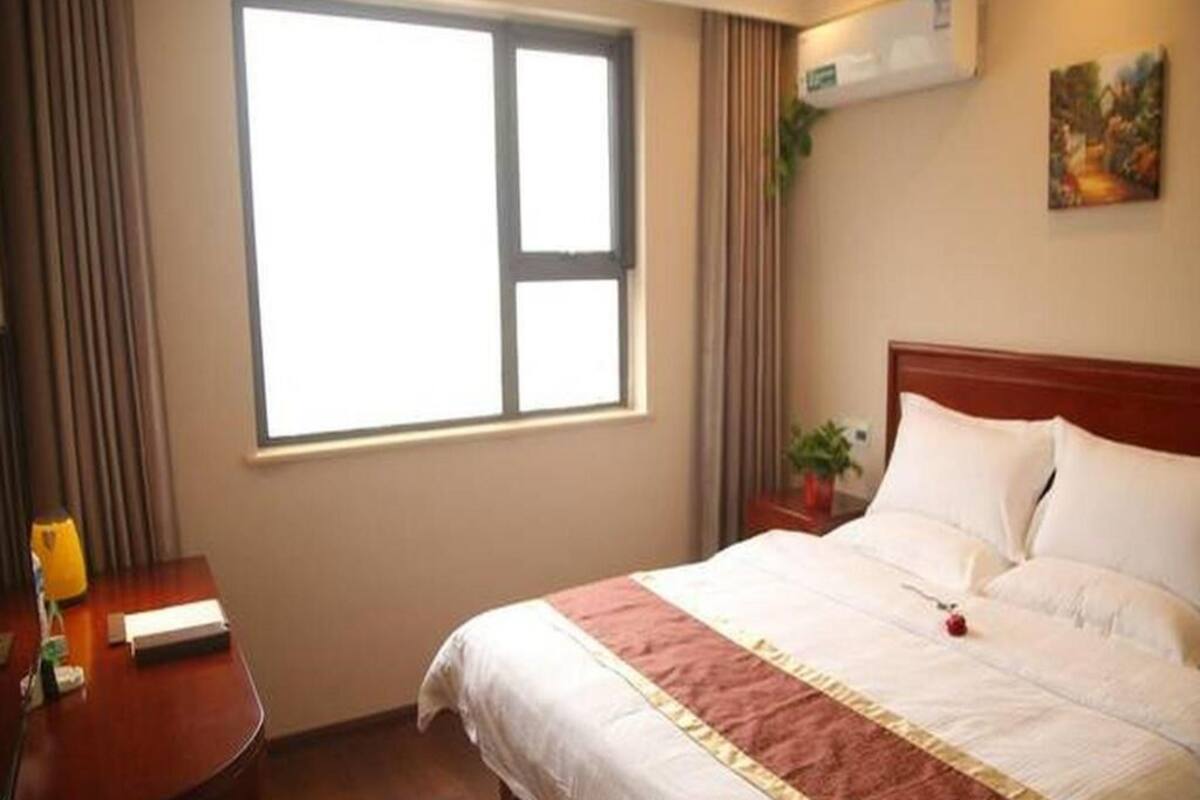 Ein Hotelzimmer mit großem Fenster, einem Bett mit weißer und roter Bettwäsche, einem Nachttisch mit Lampe und einer Wand-Klimaanlage.