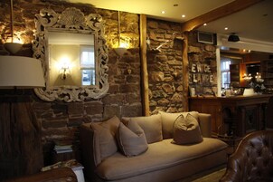 Una habitación acogedora con una pared de piedra, un espejo grande y ornamentado, un sofá con cojines y una zona de bar al fondo.