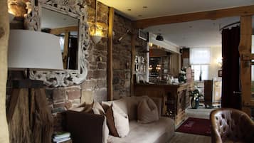 A cozy living room with a stone wall, a wooden beam ceiling, a sofa, a leather armchair, and a bar area in the background.