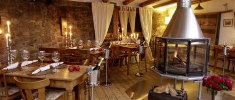 A rustic dining area with wooden tables and chairs, candles, and a central wood-burning stove.
