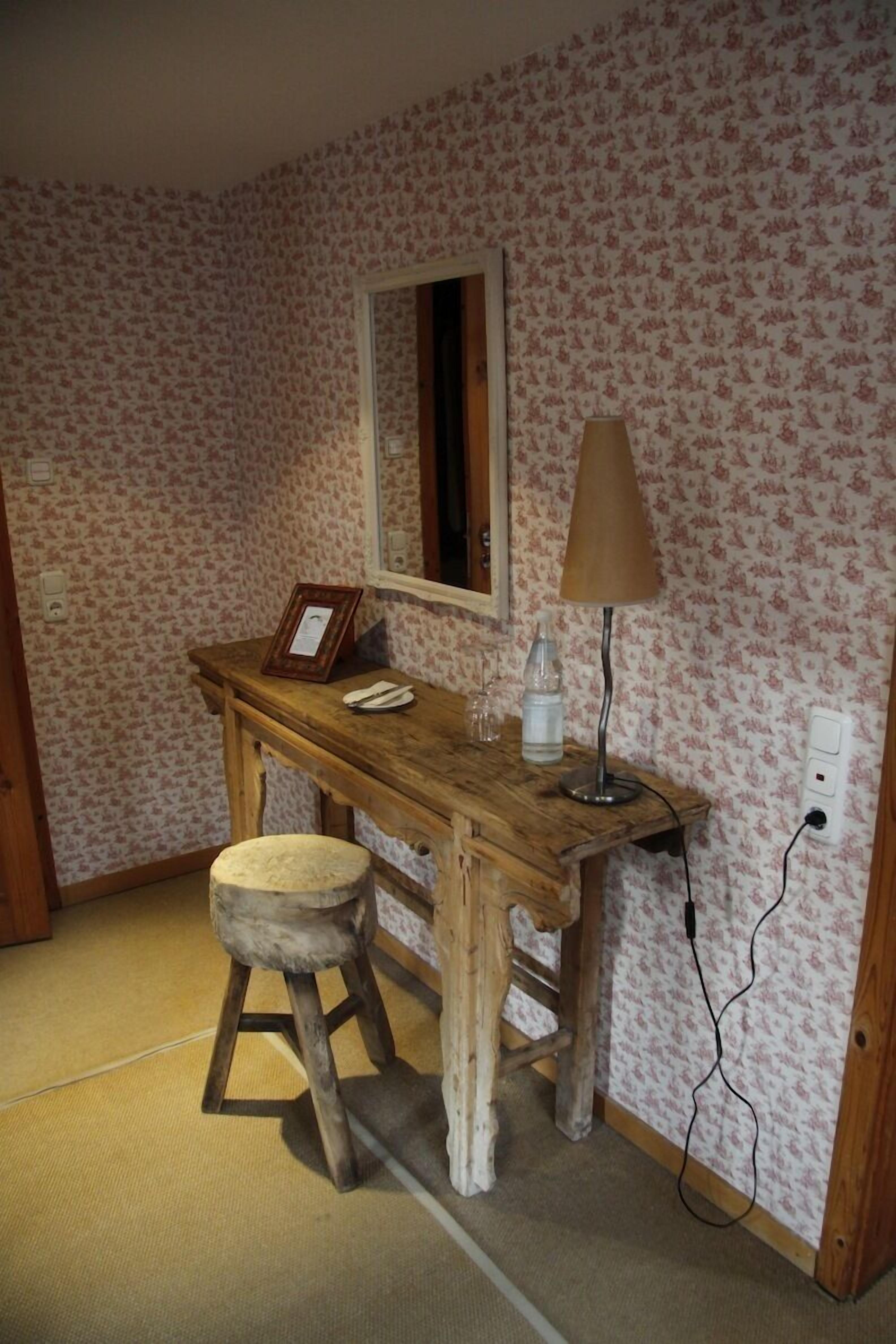 Una habitación con un escritorio de madera, un taburete, un espejo, una lámpara y un cuadro enmarcado.