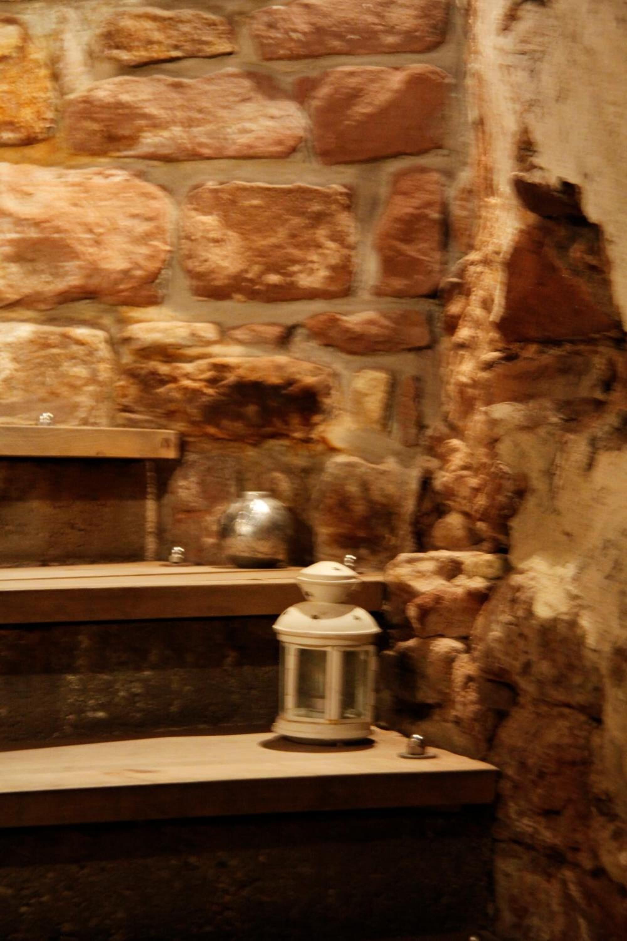 A stone wall with a rustic appearance, a shelf with a metal pot, and a lantern on a ledge.