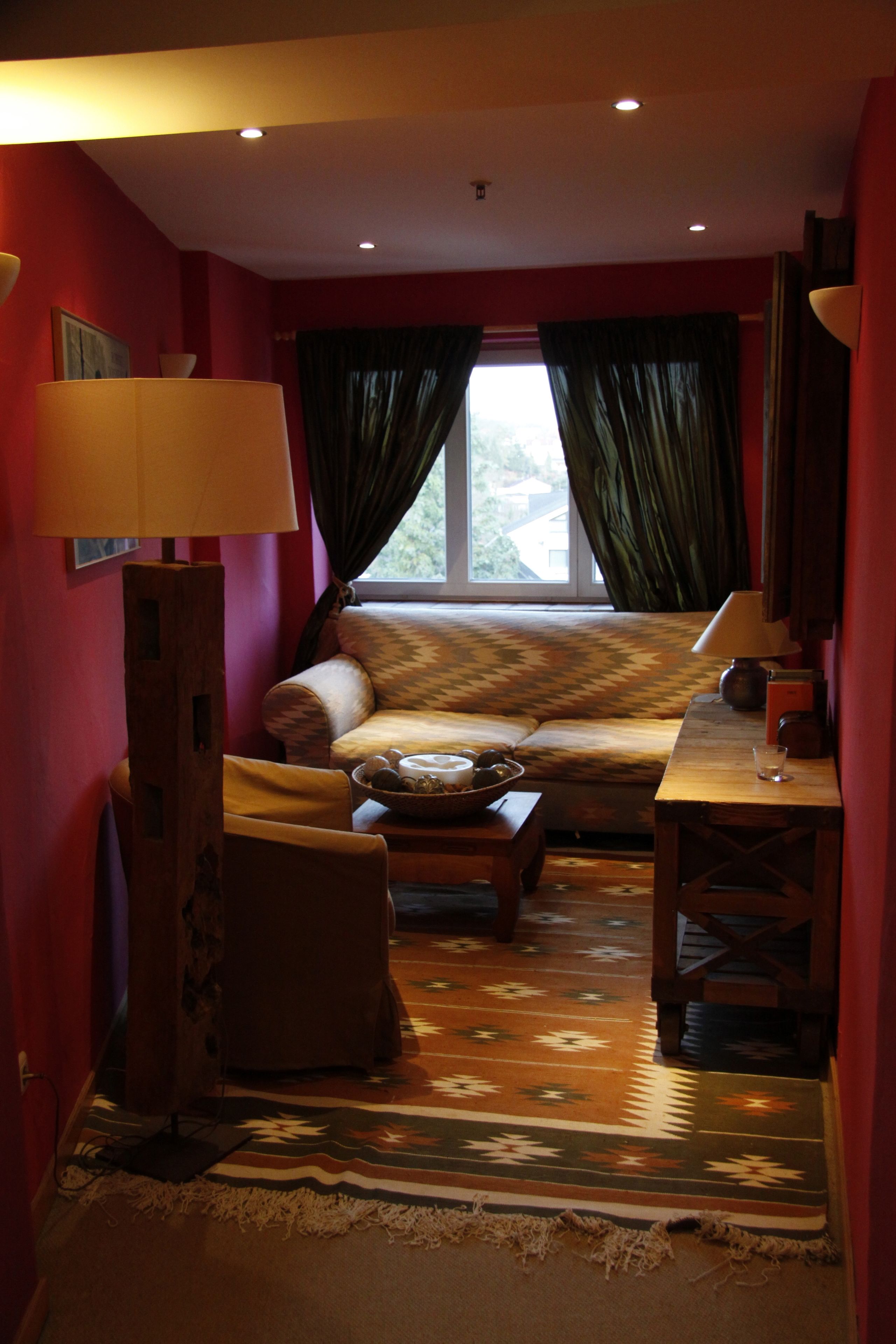 A cozy room with a sofa, a small table, and a lamp. There is a window with curtains, and the room has a warm color scheme.