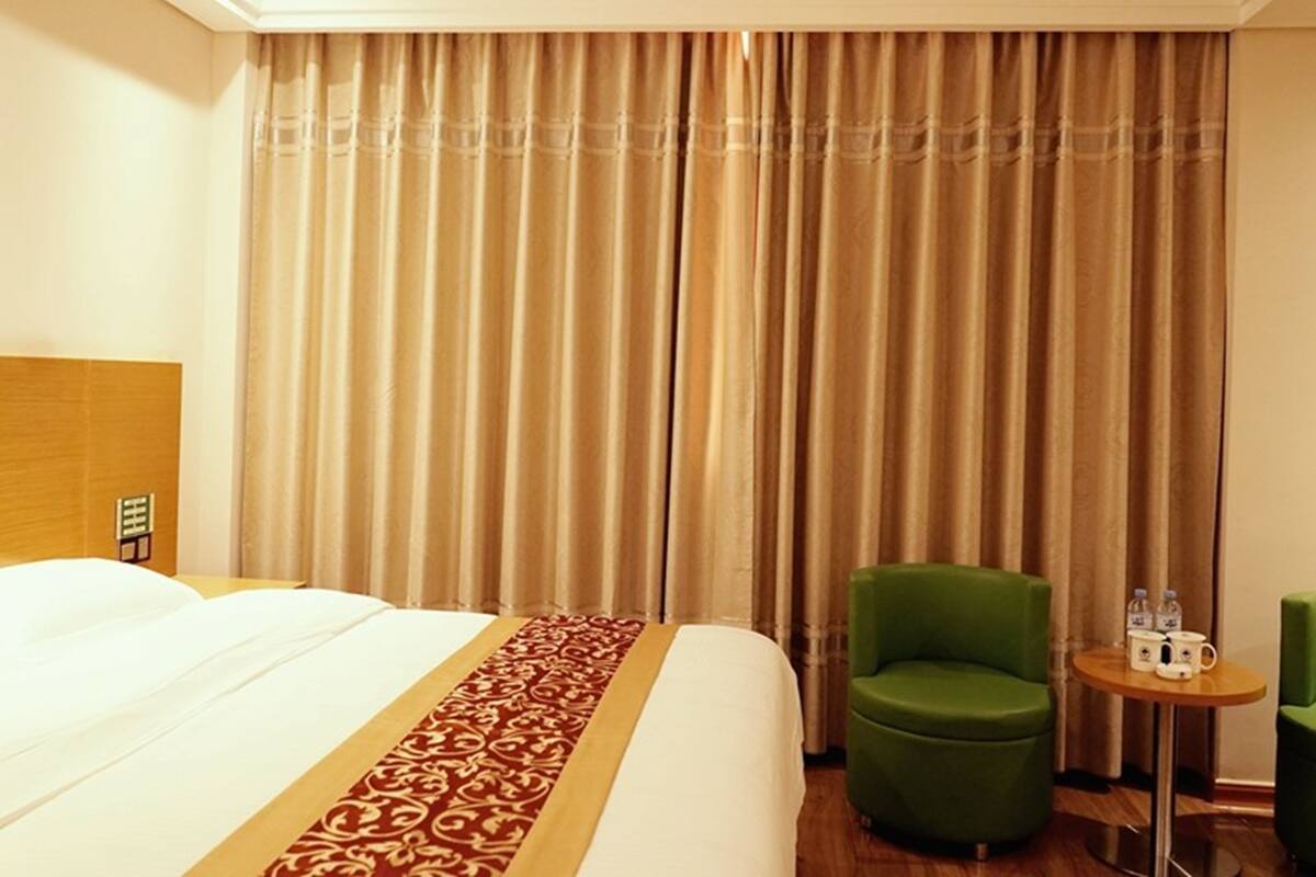 A hotel room with a bed, a green armchair, a small round table with a water bottle and a mug, and a curtain with a vertical pattern.