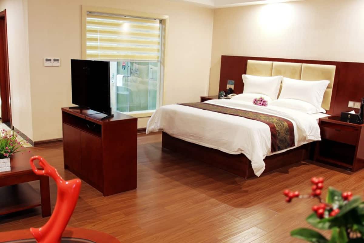 A hotel room with a large bed, a desk with a television, and a window with blinds.