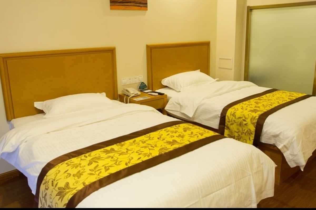 Habitación de hotel con dos camas individuales, cada una con cabecero de madera y ropa de cama blanca. Hay una mesita de noche con un teléfono y una pequeña mesa con lámpara.