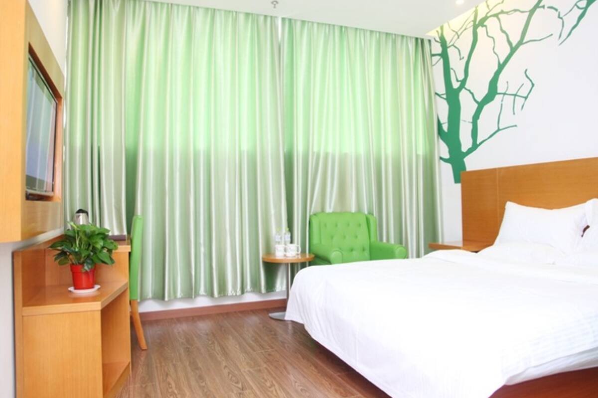 Une chambre d’hôtel avec un lit, un fauteuil vert, une petite table avec une plante et une fenêtre avec des rideaux verts.
