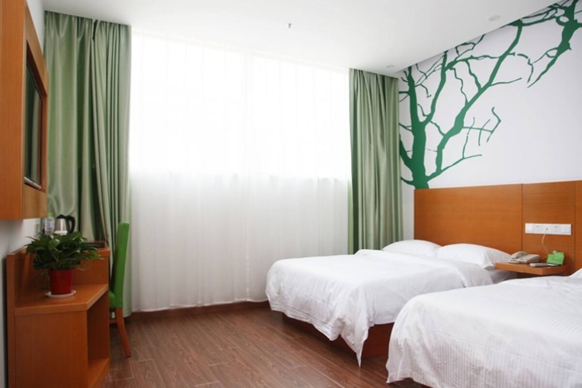 Une chambre d’hôtel avec deux lits, une table de chevet en bois ornée d’une plante, un téléphone fixé au mur et une fresque représentant un arbre vert.