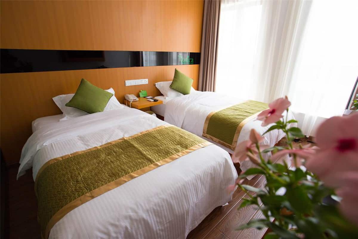 A hotel room with two beds, each with a green pillow and a white bedspread. There is a wooden headboard, a nightstand with a phone and a lamp, and a window with curtains.