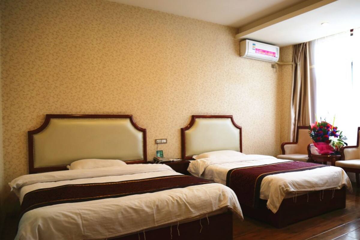 A hotel room with two beds, a wooden floor, a wall-mounted air conditioner, and a window with curtains.