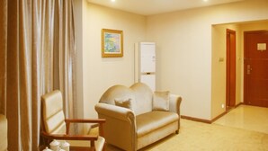 Une chambre d’hôtel avec un canapé beige, une chaise en bois, un tableau accroché au mur et une porte donnant sur une autre pièce.