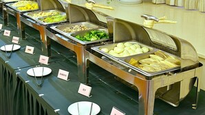 Un bufé con diversos alimentos en recipientes metálicos, cada uno identificado con una etiqueta.