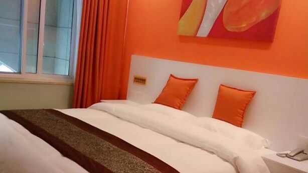 Een hotelkamer met een bed, oranje gordijnen, een wit hoofdeinde en een schilderij aan de muur.