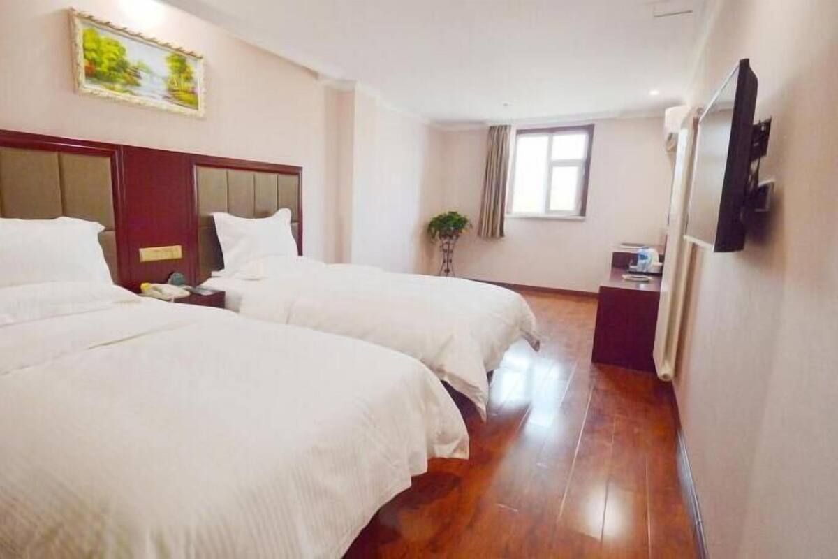 Een hotelkamer met twee bedden, een houten hoofdbord, een klein nachtkastje met een lamp, een raam met gordijnen en een televisie aan de muur.