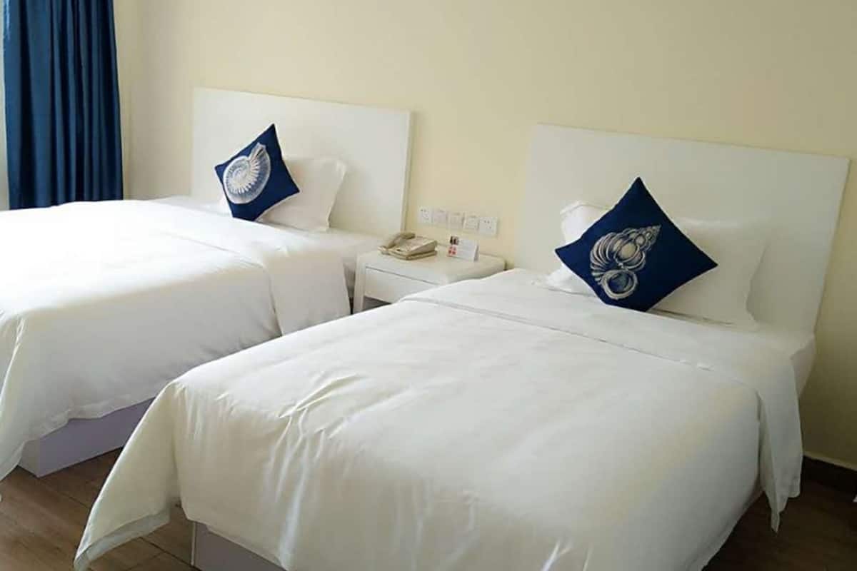 A hotel room with two single beds, each with a white bedspread and a blue pillow. There is a bedside table with a phone and a small lamp.