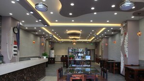 Un restaurante con zona de comedor central, varias mesas y sillas. Hay luces de techo y una recepción con mostrador.