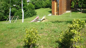 A modern wooden building with a sloped roof, surrounded by lush greenery and a playground. - Hotel Residence La Rosa (Castione della Presolana)