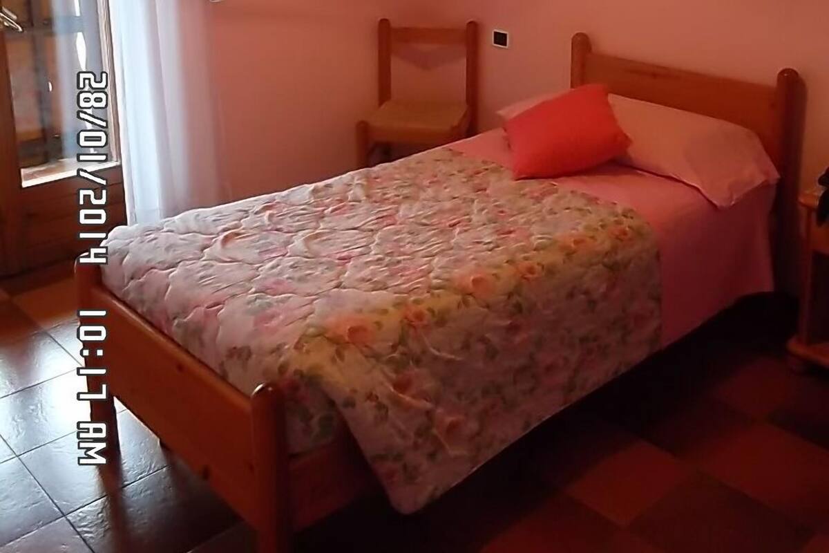 A bedroom with a wooden bed, floral bedspread, a red pillow, a wooden chair, and a painting on the wall.
