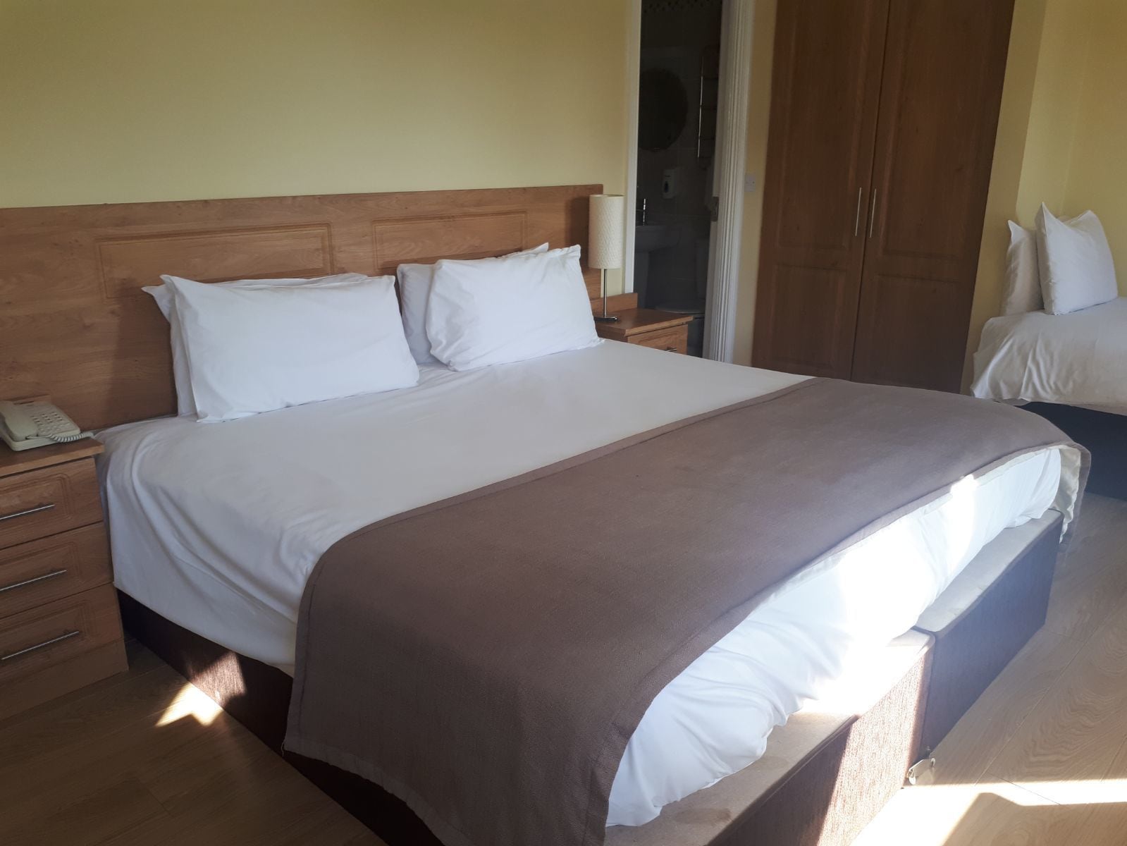 Een net opgemaakt bed met witte lakens en een bruine plaid, een houten nachtkastje met een telefoon, en een houten garderobe in een hotelkamer.