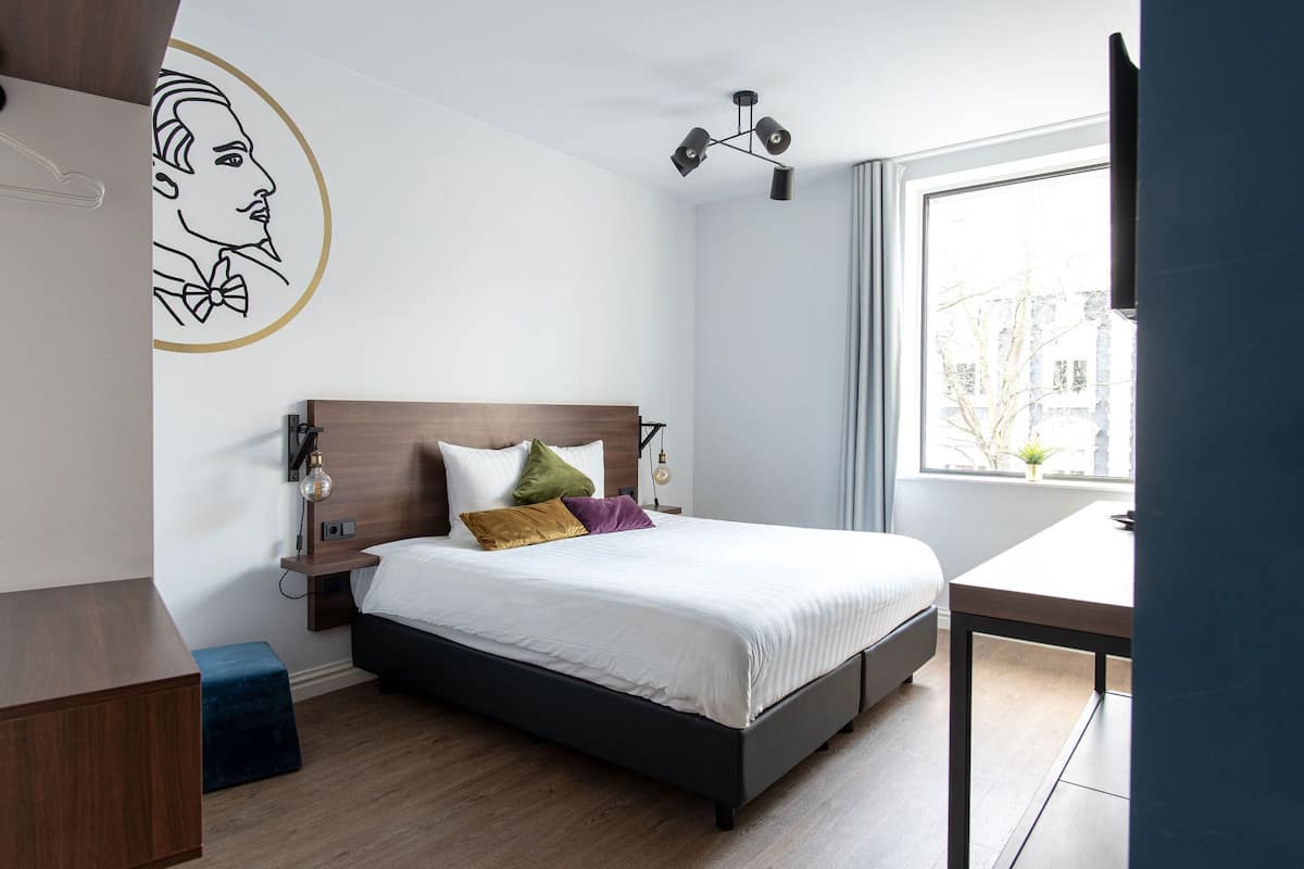A modern bedroom with a wooden headboard, a large window, a bedside table, and a wall art piece.