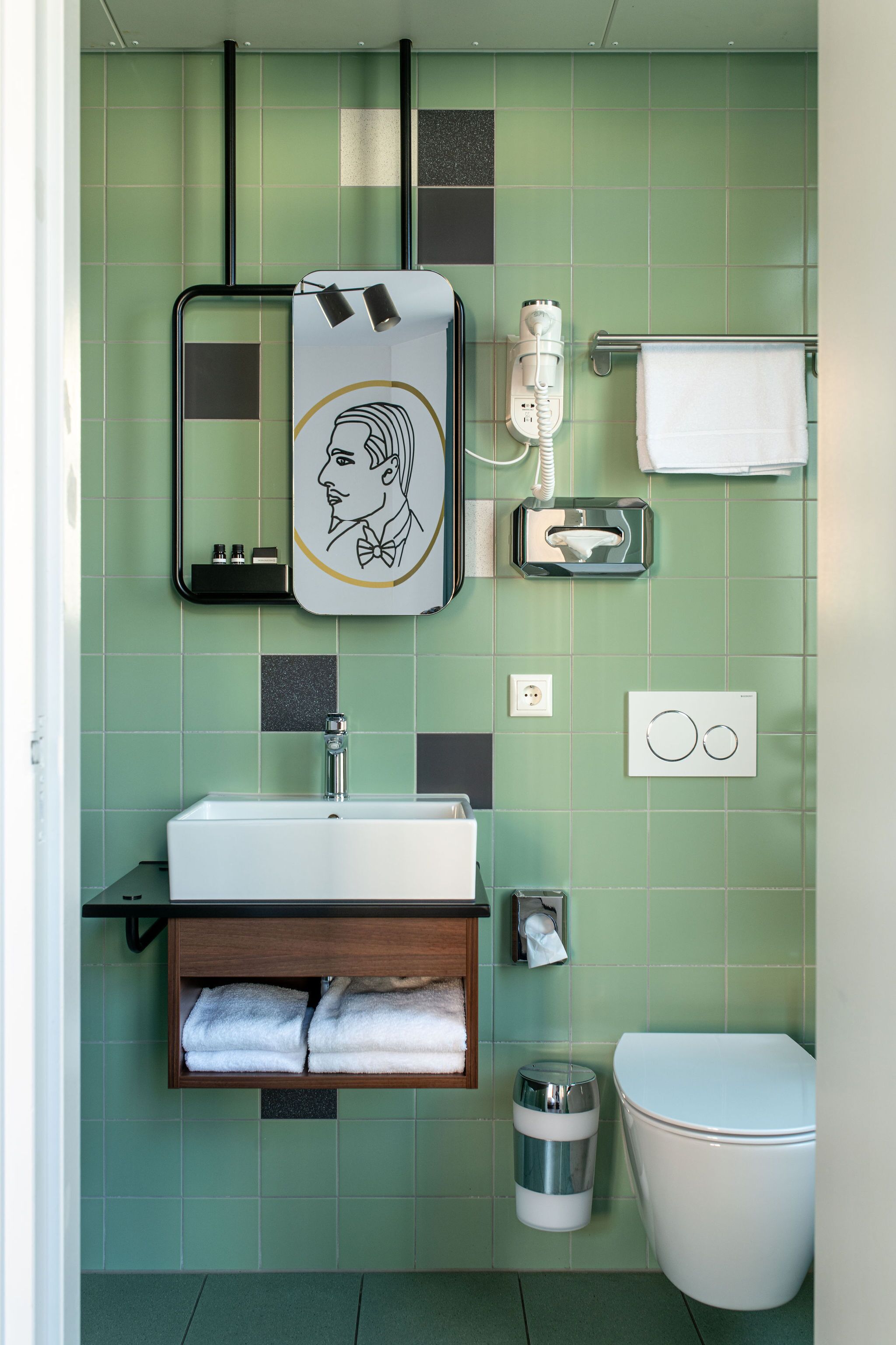 Un baño con pared de azulejos verdes, un lavamanos con espejo, toallero, inodoro y cubo de basura.