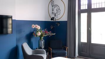Habitación de hotel con cama, dos sillones, un jarrón con flores, televisor montado en la pared y un ventanal con cortinas.