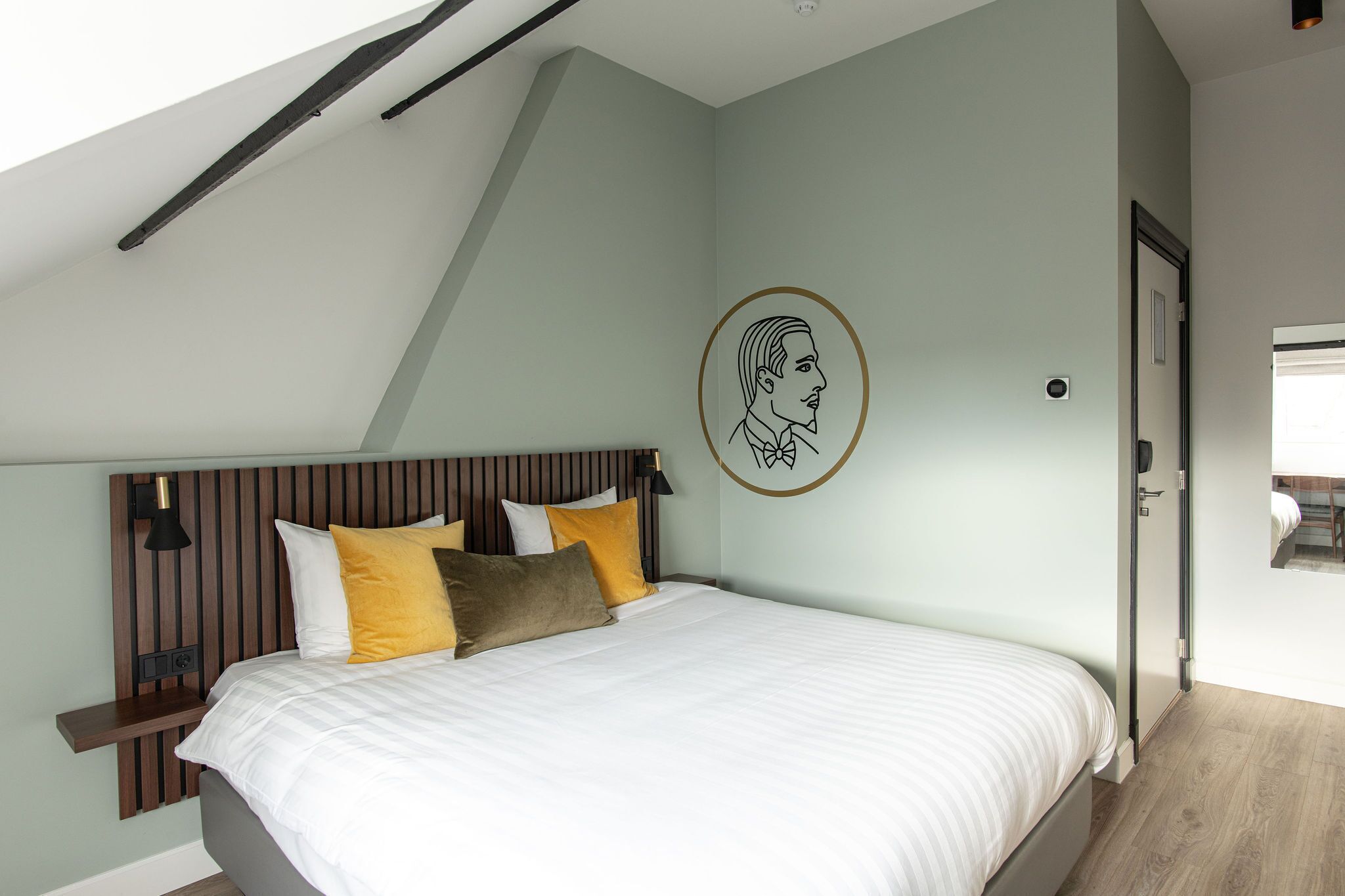 A bedroom with a sloped ceiling, a bed with white bedding and multiple pillows, a wall-mounted light fixture, and a door with a mirror.