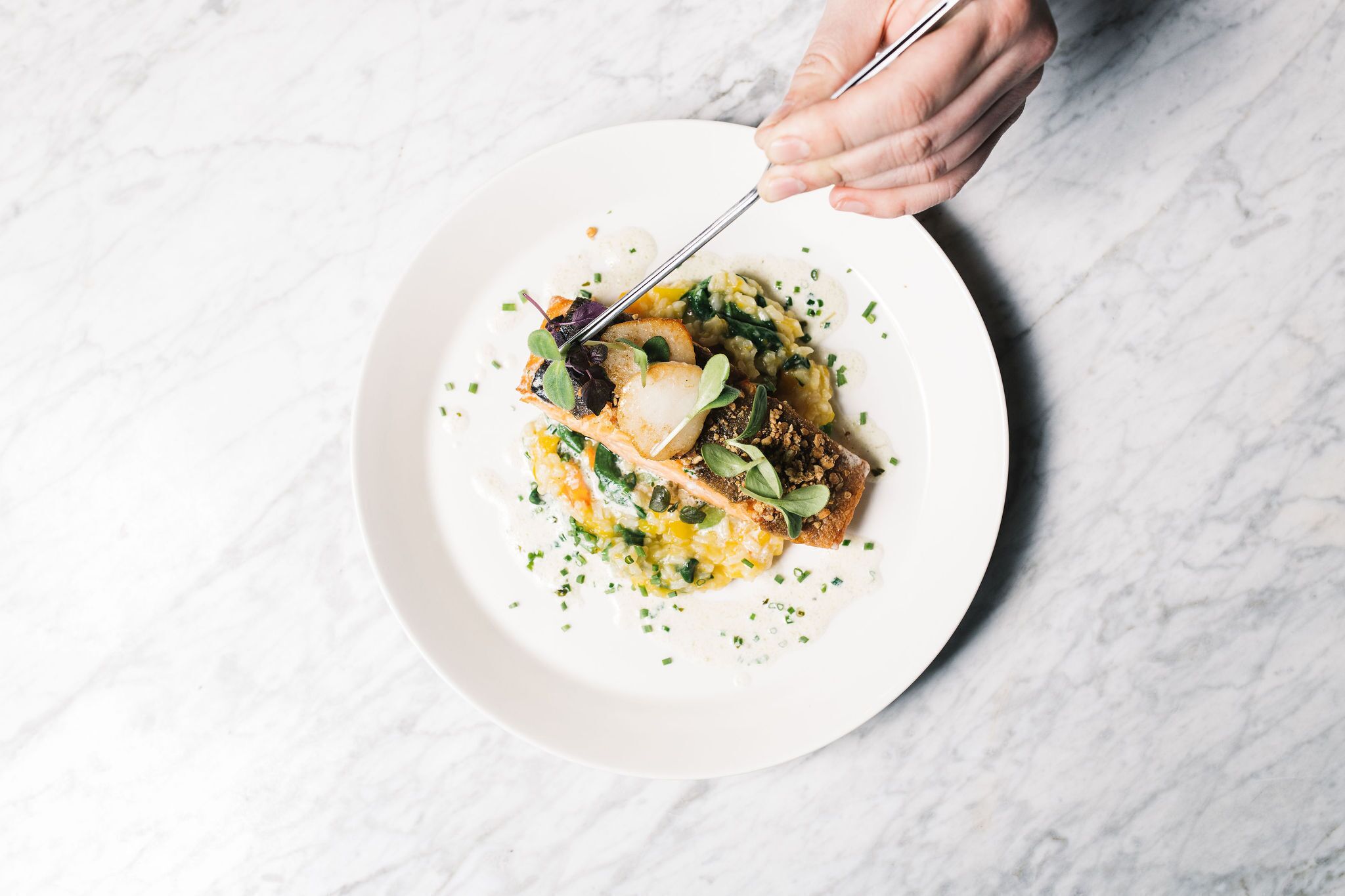 A white plate with a dish of food, including fish, greens, and possibly some grains, on a marble surface.