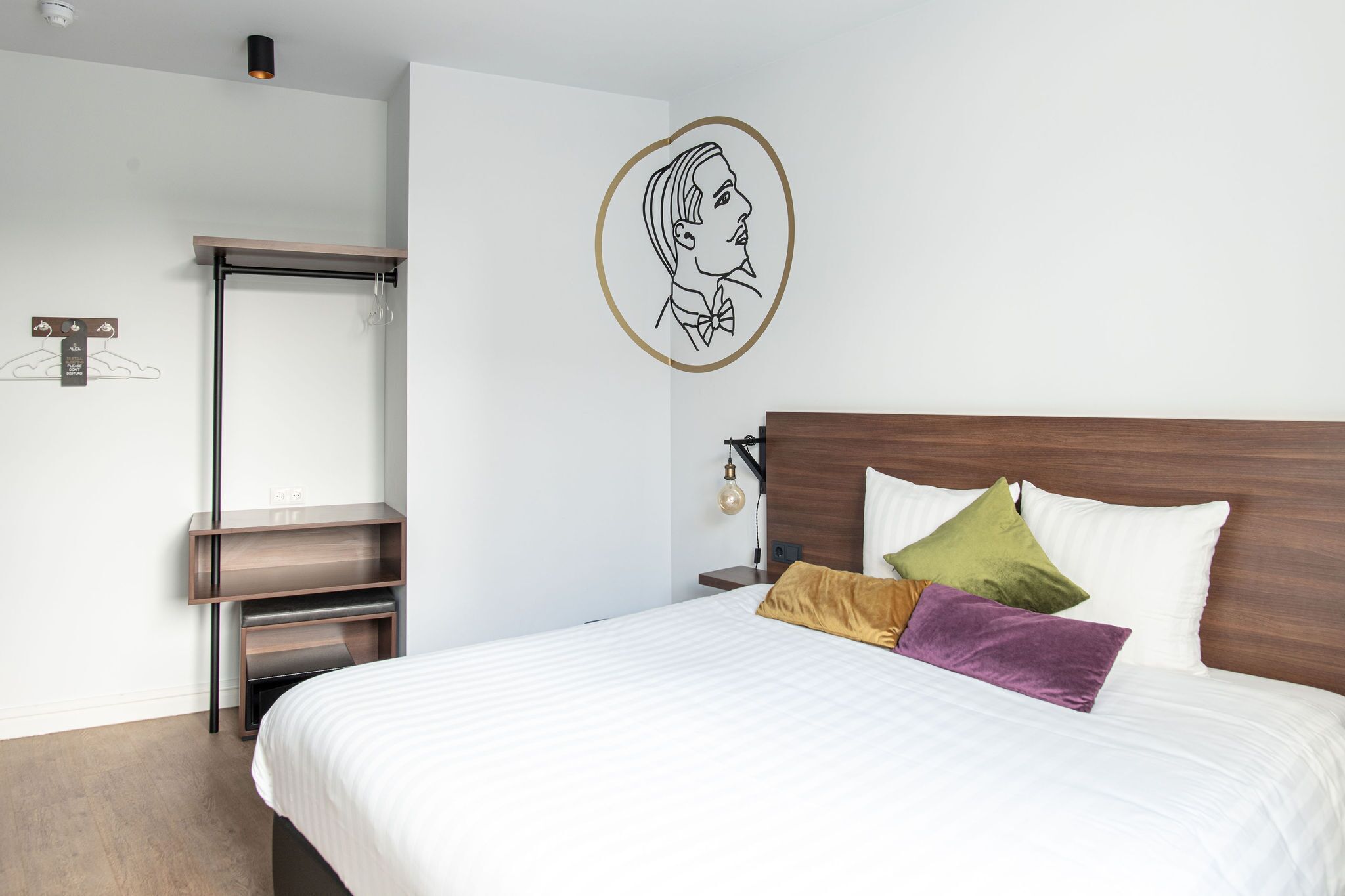 A neatly made bed with white bedding and multiple pillows, a wooden headboard, a wall-mounted light fixture, and a coat rack.